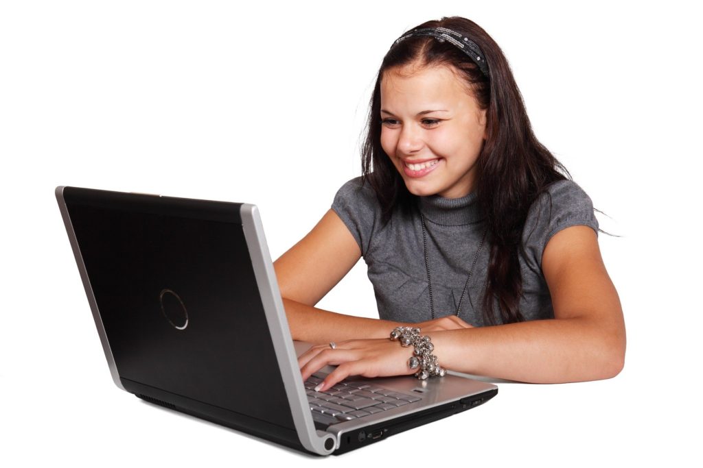 Zdjęcie przedstawia uśmiechniętą dziewczynę siedzącą przed otwartym laptopem.