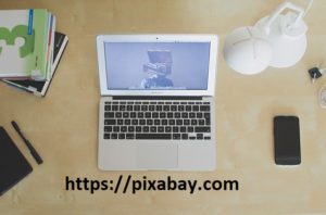 otwarty laptop na biurku, po prawej stronie laptopa telefon komórkowy, po lewej książki i długopis. Poniżej laptopa link do strony pixabay: https://pixabay.com